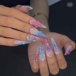 May include: A hand with long, pointed acrylic nails. The nails feature a futuristic design with iridescent colors, including pink, blue, and purple. Some nails have 3D embellishments and metallic accents, creating a space-age aesthetic.