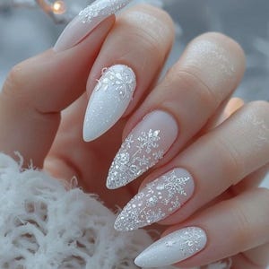 May include: Close-up of a hand with long, pointed, white and glittery nail art. Some nails are solid white, while others feature a sheer base with silver snowflake designs. The nails are a winter-themed manicure.