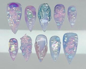 May include: A set of ten artificial nails in various shapes and designs. The nails feature a pearlescent, iridescent finish with a mix of light blue, purple, and pink hues. Some nails have textured, bubble-like details, while others showcase abstract patterns.