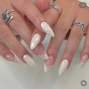May include: A close-up of a hand with long, almond-shaped nails. The nails feature a variety of designs, including white with pink dots, clear with white bows, and a French tip design with pink accents. Silver rings adorn the fingers.