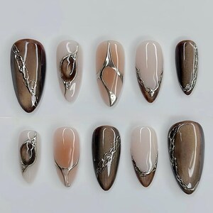 May include: A collection of ten almond-shaped false nails. The nails display diverse designs, including solid brown, nude, and ombre effects. Each nail is decorated with silver metallic accents, creating a modern and elegant appearance.