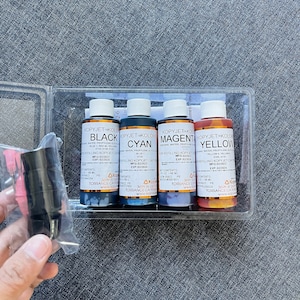 May include: A clear plastic case containing four bottles of Kopyjet Kolors ink in black, cyan, magenta, and yellow. The bottles are labeled with the ink color and have a capacity of 60 ml. Also included are black ink cartridges and pink needles.