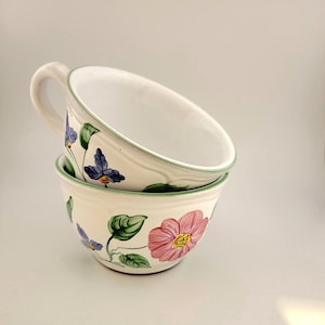 May include: Two white ceramic bowls stacked, one with a handle. Each bowl is decorated with a floral pattern featuring pink and purple flowers, green leaves, and a green rim. The bowls are ideal for serving soup or cereal.