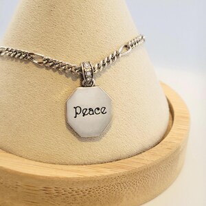 K&ouml;nnte beinhalten: Silberfarbene Halskette mit Kette und Anh&auml;nger. Der Anh&auml;nger hat eine achteckige Form mit dem Wort "Peace" in Schreibschrift. Die Oberseite des Anh&auml;ngers hat kleine, klare Steine. Die Halskette wird auf einem hellbraunen, kegelf&ouml;rmigen St&auml;nder pr&auml;sentiert.