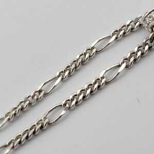 K&ouml;nnte beinhalten: Nahaufnahme von zwei silbernen Figaro-Ketten. Die Ketten haben ein sich wiederholendes Muster aus drei kleinen kreisf&ouml;rmigen Gliedern, gefolgt von einem l&auml;nglichen ovalen Glied. Die Ketten werden auf wei&szlig;em Hintergrund dargestellt.