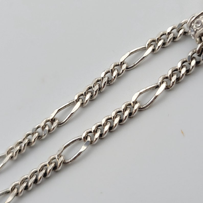 K&ouml;nnte beinhalten: Nahaufnahme von zwei silbernen Figaro-Ketten. Die Ketten haben ein sich wiederholendes Muster aus drei kleinen kreisf&ouml;rmigen Gliedern, gefolgt von einem l&auml;nglichen ovalen Glied. Die Ketten werden auf wei&szlig;em Hintergrund dargestellt.