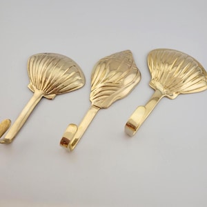 May include: Three polished gold-toned metal wall hooks, each featuring a distinct seashell design at the top. The hooks have a smooth, reflective surface and a curved end for hanging items. The designs include two fan-shaped scallop shells and one spiral conch shell, all arranged in a row on a plain surface.