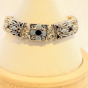 May include: Silver-toned bracelet with a floral design. The bracelet features light blue and black enamel accents. The central flower is square-shaped, and the bracelet is displayed on a white surface.