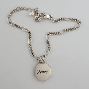 K&ouml;nnte beinhalten: Silberfarbenes Armband mit Kettenglied-Design. Das Armband hat eine kleine Perle, einen herzf&ouml;rmigen Verschluss und einen achteckigen Anh&auml;nger mit der Aufschrift "Peace". Der Anh&auml;nger ist mit kleinen, klaren Kristallen verziert.