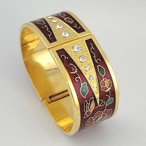 May include: A gold-tone hinged bangle bracelet featuring a deep burgundy enamel finish with floral motifs in green, pink, and blue. The center of the bracelet displays a triangular gold panel inlaid with four clear, round-cut rhinestones.