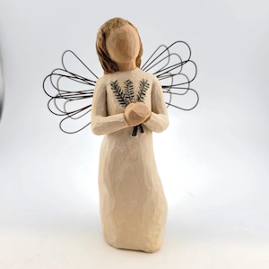 May include: A carved wooden angel figurine with a cream-colored dress and brown hair. The angel holds a bundle of green sprigs and has wire wings. The figurine is approximately 6 inches tall.