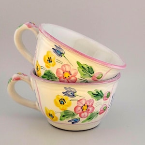 May include: Two stacked ceramic teacups with floral designs. The cups are cream-colored with pink rims and handles. The floral pattern includes pink, yellow, and blue flowers with green leaves. The cups are stacked at a slight angle.