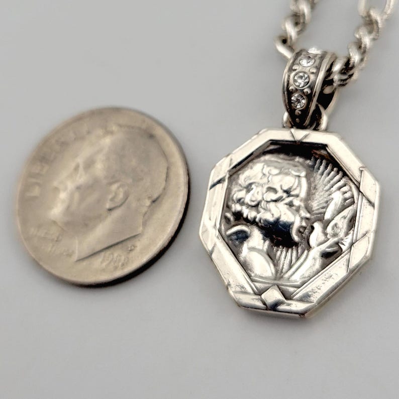 K&ouml;nnte beinhalten: Eine silberne Anh&auml;ngerkette mit einem achteckigen Anh&auml;nger mit einem detaillierten Relief einer klassischen Figur. Der Anh&auml;nger ist an einer Silberkette befestigt. Der Anh&auml;nger befindet sich neben einer US-Zehn-Cent-M&uuml;nze.