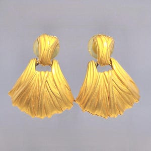 May include: A pair of gold-colored earrings. Each earring features a fan-shaped design with textured lines, attached to a smaller, rounded top. The earrings have a bright, metallic finish, reflecting light. The overall design is elegant and detailed.