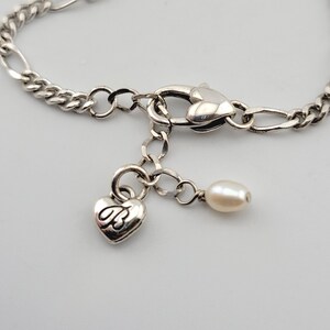 K&ouml;nnte beinhalten: Silberfarbenes Armband mit herzf&ouml;rmigem Anh&auml;nger und kleinem Perlenanh&auml;nger. Der Anh&auml;nger hat ein kursives "B". Das Armband hat einen Karabinerverschluss und ein Kettenglied-Design.