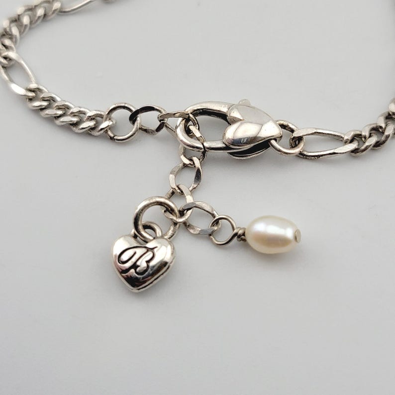 K&ouml;nnte beinhalten: Silberfarbenes Armband mit herzf&ouml;rmigem Anh&auml;nger und kleinem Perlenanh&auml;nger. Der Anh&auml;nger hat ein kursives "B". Das Armband hat einen Karabinerverschluss und ein Kettenglied-Design.