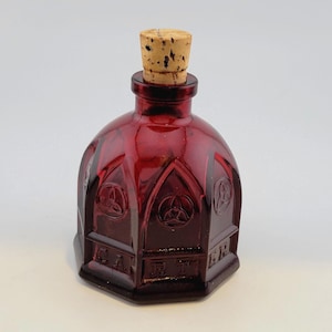 May include: A deep red glass bottle with a cork stopper. The bottle features a faceted, gothic-inspired design with embossed circular symbols and the text CARTER molded into the base panels. The glass is translucent and dark in color.