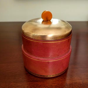 May include: A two-tiered, round red box with a gold-colored lid and a small orange knob on top. The box has gold trim and sits on a dark wooden surface. The box appears to be vintage.
