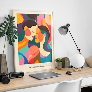 Puede incluir: Impresión artística enmarcada con un diseño abstracto colorido de dos personas besándose. La obra presenta tonos vibrantes de naranja, rosa, azul y verde. El texto "Too cool to be binary" está impreso en la parte inferior. En un escritorio de madera con una lámpara.