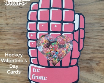 May include: A pink hockey glove Valentine's Day card with a heart-shaped window filled with colorful candies. The card has the text "to:" and "from:" on the bottom. The card is on a wooden surface.