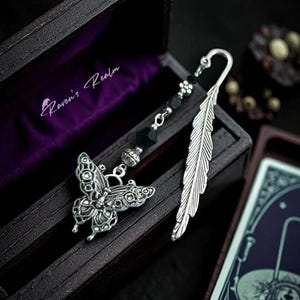May include: A silver bookmark featuring a butterfly and feather design, presented in a dark wooden box with a purple velvet interior. The bookmark includes black beads and the text "Raven's Realm".