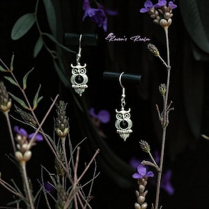 Könnte beinhalten: Silberne Eulenohrringe mit schwarzen Perlenaugen, vor einem dunklen Hintergrund mit lila Blumen und Blättern. Die Ohrringe weisen ein detailliertes Eulen-Design auf, das eine Vintage- oder Gothic-Ästhetik suggeriert. Der Text "Raven's Realm" ist sichtbar.