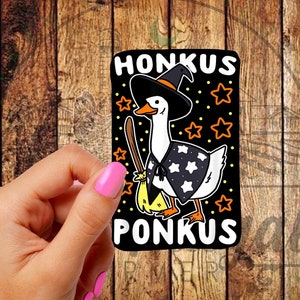 May include: A black sticker featuring a cartoon goose dressed as a witch, holding a broom. The goose wears a black hat and cape with star patterns. The text "HONKUS PONKUS" is displayed above and below the goose.