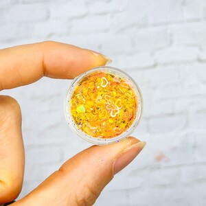 May include: A small clear container filled with yellow, orange, and white glitter. The glitter is in a variety of shapes and sizes.