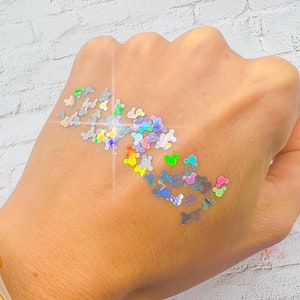 May include: A hand with iridescent holographic glitter in the shape of Mickey Mouse heads. The glitter is scattered on the back of the hand, creating a sparkly effect.