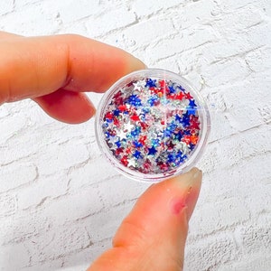 May include: A small, clear container filled with star-shaped glitter in red, white, and blue. The container is held between fingers against a white brick background. The glitter is a mix of sizes and reflects light.