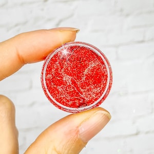 May include: A small clear container filled with red glitter. The glitter is a fine, sparkly texture.