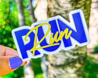 以下が含まれることがあります： スタイリッシュなフォントで「Run」と書かれた青と黄色のステッカー。ステッカーは、木々のぼやけた背景の前で、手で持たれています。