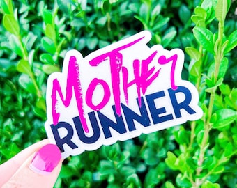 May include: A white sticker with the words "Mother Runner" in pink and blue lettering. The sticker is held in a hand against a green leafy background.
