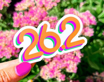 May include: A sticker with the number 26.2 in a retro font with orange, green, and purple outlines. The sticker is held in a hand against a background of pink flowers.