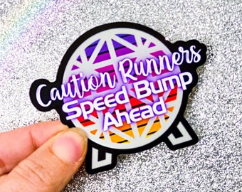 Puede incluir: Una pegatina en blanco y negro con un gráfico colorido de una esfera con rayas. El texto "Caution Runners Speed Bump Ahead" está escrito en morado en la pegatina.