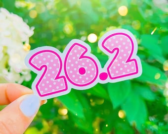 May include: Pink and white polka dot sticker with the number 26.2 on it. The sticker is held in a hand against a green leafy background.