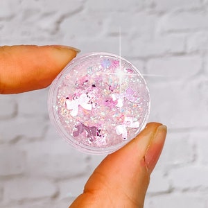 May include: A small clear jar filled with pink and white glitter and iridescent holographic glitter. The glitter is in the shape of stars and hearts.