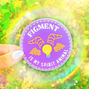 Puede incluir: Una pegatina redonda morada con el texto "FIGMENT IS MY SPIRIT ANIMAL" en blanco. La pegatina presenta una bombilla amarilla con alas y dise&ntilde;os de alas amarillas. La pegatina est&aacute; sobre un fondo verde y amarillo borroso.