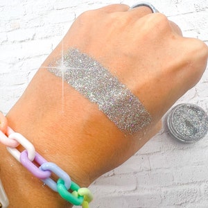 May include: A silver glitter on a person's hand with a silver glitter jar in the background.