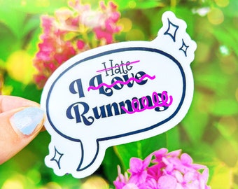 May include: A white sticker with a speech bubble shape. The sticker says "I love running" with the word "hate" crossed out and the word "love" underlined. The sticker is held by a hand against a background of pink flowers and green foliage.