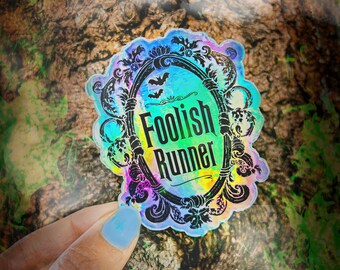 May include: A holographic sticker with a black ornate frame and the text "Foolish Runner" in black lettering. The sticker has a rainbow iridescent finish.