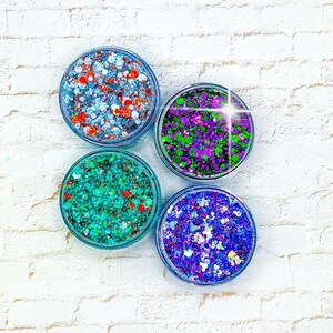 May include: Four small jars of glitter, each containing a different color combination. The jars are arranged in a circle on a white brick background. The glitter colors are blue, green, purple, and orange.