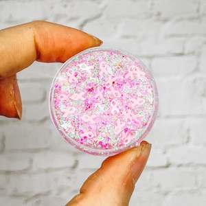 May include: A small clear jar filled with pink glitter and pink ribbon charms. The jar is being held in a hand.