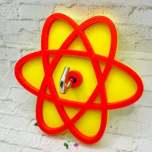 May include: A red and yellow wall decor piece in the shape of an atom. The atom has a yellow center with a red outline and a silver hook. The decor is mounted on a white brick wall.