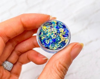 May include: A small clear jar filled with blue, green, and yellow glitter. The glitter is in the shape of stars and small pieces of confetti.