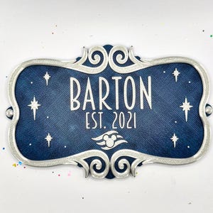 May include: A rectangular navy blue sign with a silver scrollwork border. The text reads BARTON EST. 2021 in white lettering. A white graphic of a stylized cruise ship icon with a mouse head silhouette is centered below the text, surrounded by white star accents.