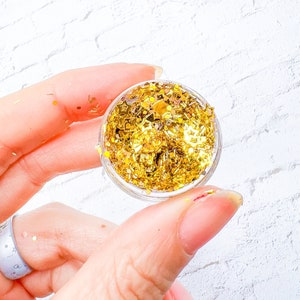 May include: A small, clear container filled with gold glitter. The glitter includes star and irregular shapes. The container is held between two fingers against a white brick background. The glitter is a bright, shiny gold color.