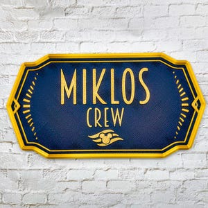 May include: A rectangular sign with a dark blue textured background and a bright yellow border. The text MIKLOS CREW is printed in yellow, accompanied by a yellow graphic of a stylized wave with a small circular icon in the center.