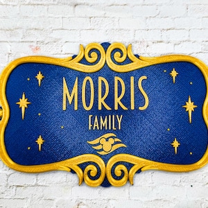 May include: A blue and gold family name sign with the name "MORRIS" and the word "FAMILY" in gold lettering. The sign has a decorative gold border and star accents. A Mickey Mouse head logo is also present.