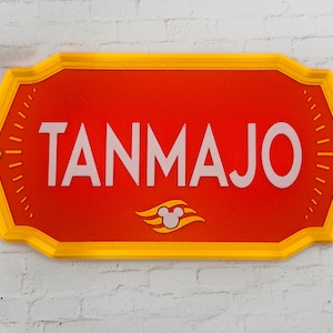 May include: A rectangular sign with the name "TANMAJO" in white capital letters on a red background. The sign has a yellow border and a Mickey Mouse logo. The sign is mounted on a white brick wall.